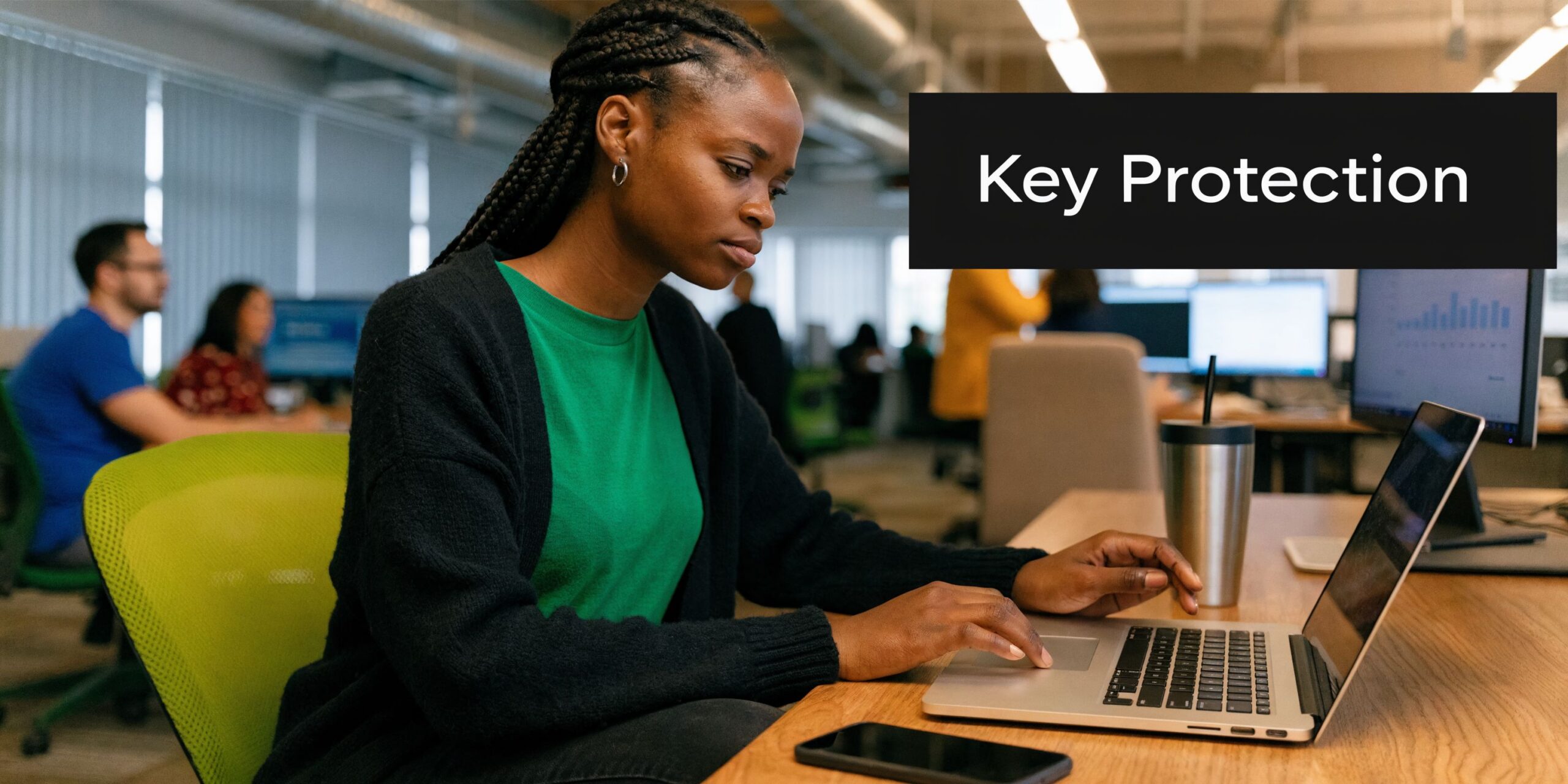 A young woman working on her laptop at an office desk with a Key Protection label nearby.