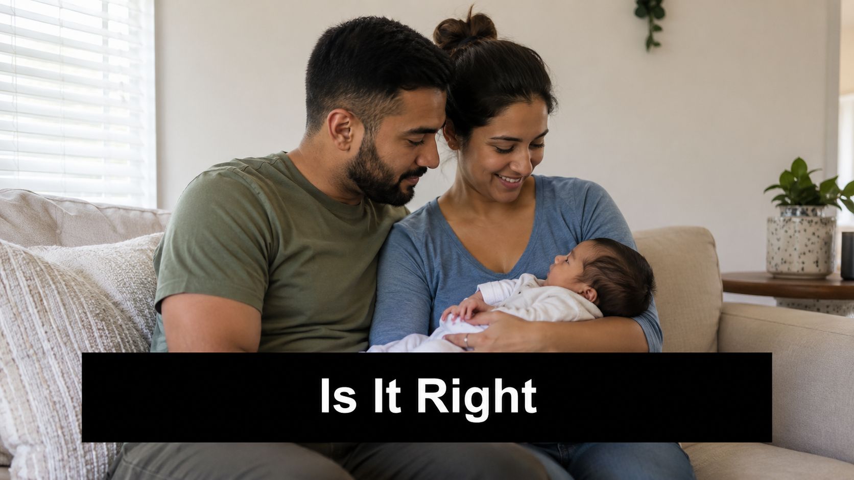 A young couple sitting together on a comfortable sofa while lovingly looking down at their newborn baby.